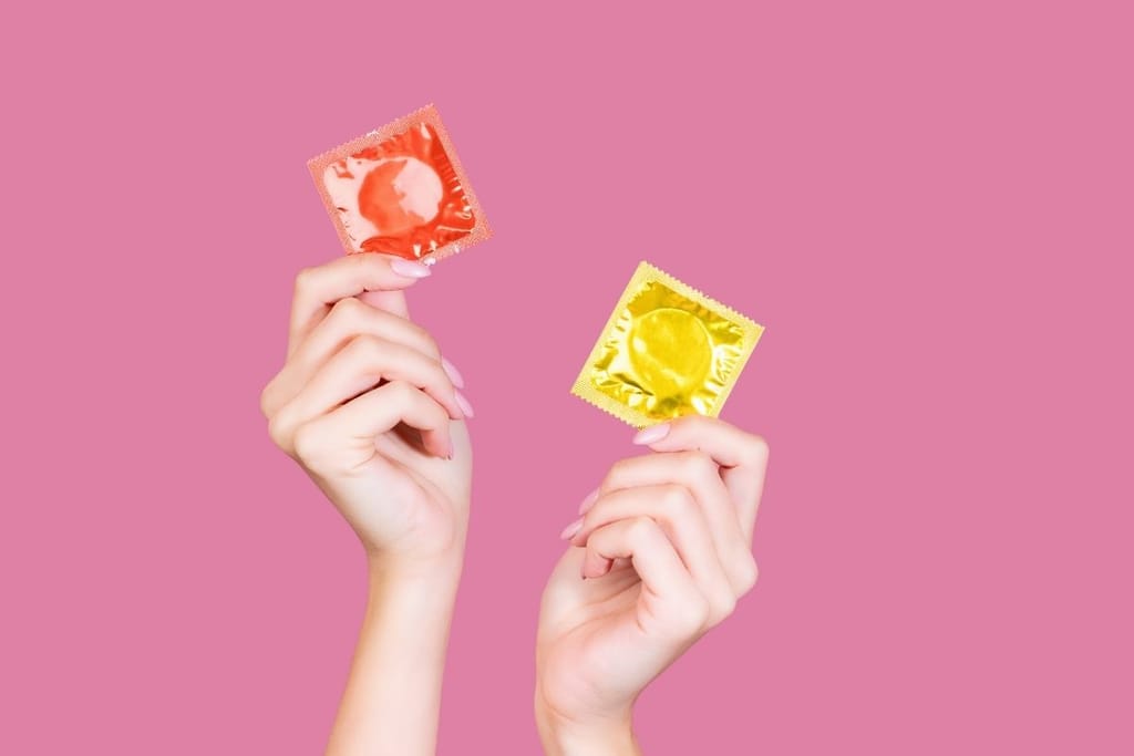 Kaksi käsissä pidettyä värikästä kondomipakkausta pinkkiä taustaa vasten – kondomien hinnat voivat nousta Iranin kriisin vuoksi