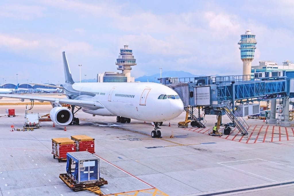 Suuri matkustajalentokone portilla lentokenttäterminaalissa, taustalla lennonjohtotornit ja kuljetuskontteja asfaltilla.