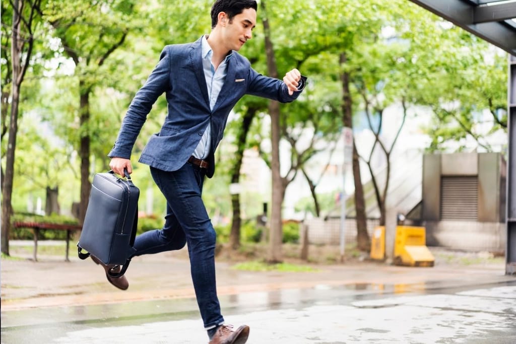 Kiireinen liikemies juoksee sateisella kadulla katsoen kelloa, myöhässä tapaamisesta