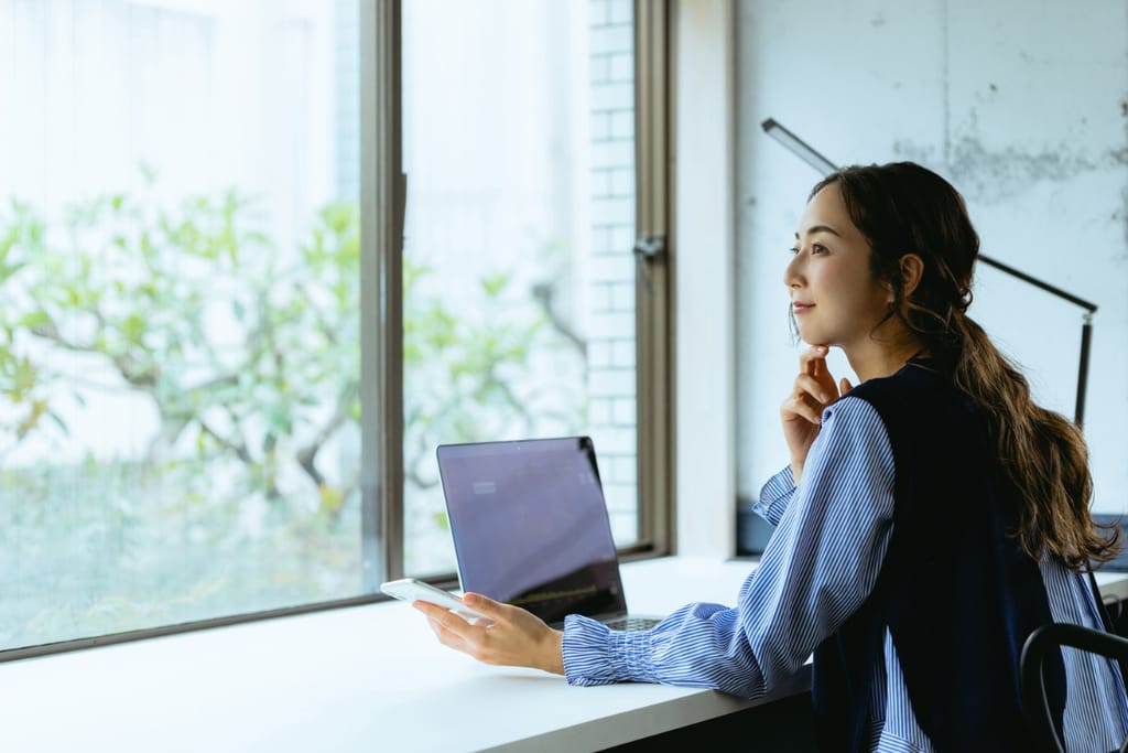 Nuori aasialaisnainen istuu tietokoneen ääressä ja katsoo ulos.