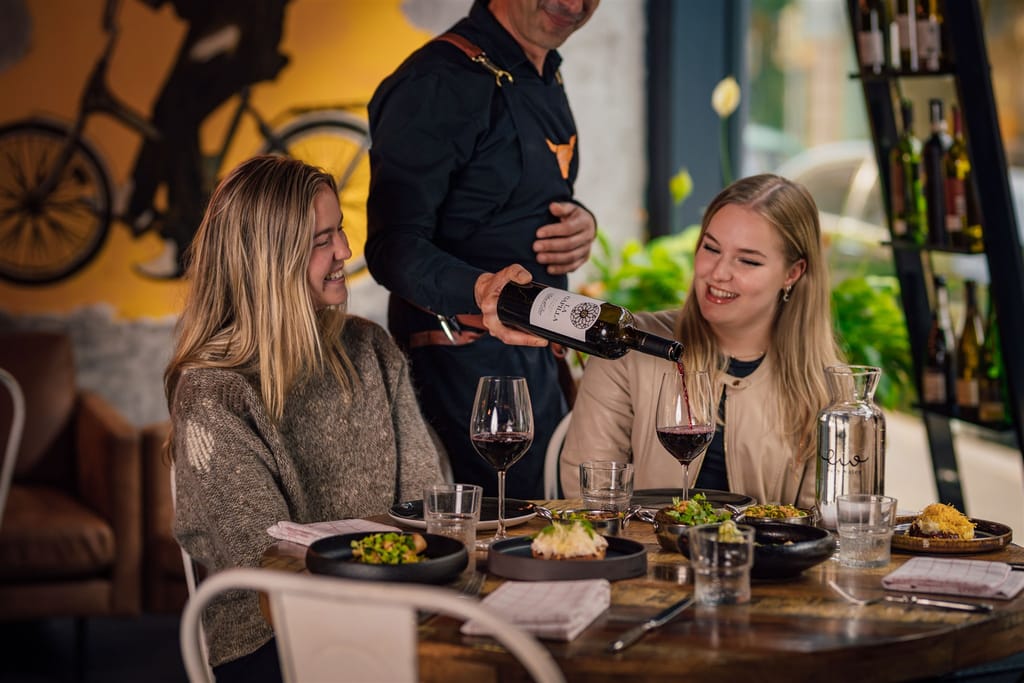 Ravintolassa kaksi naista istuu pöydässä. Pöydällä ruokia ja tarjoilija kaataa viiniä.