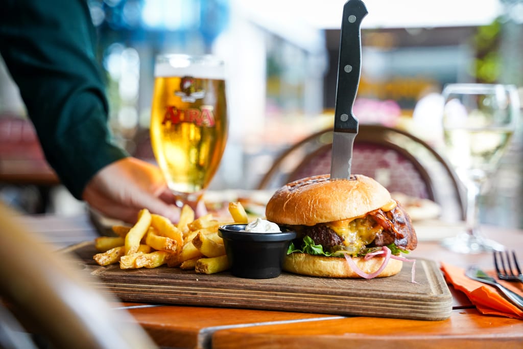 Ravintolapöydällä burgeriannos, jossa ranskalaisia ja olutlasi taustalla.