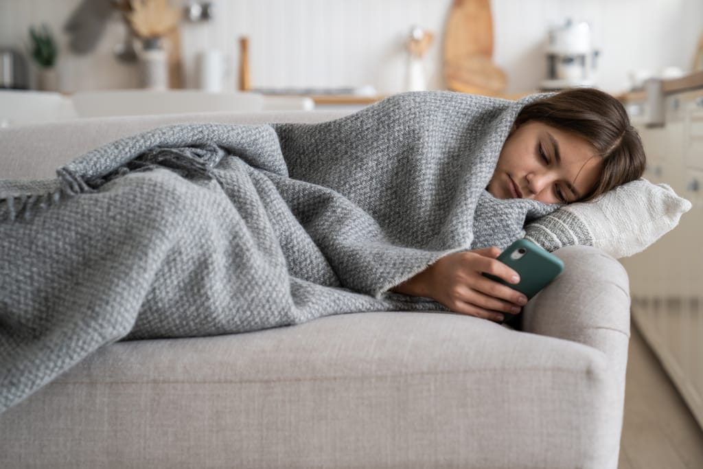 Nuori nainen makaa sohvalla viltti päällä ja selaa puhelinta.