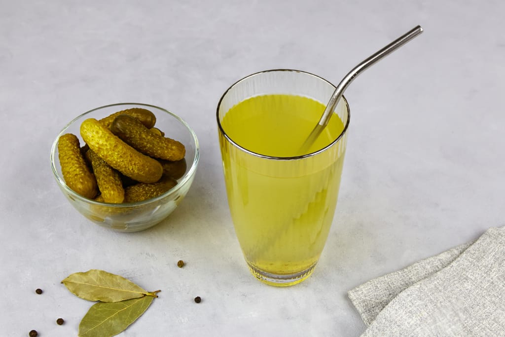 Pöydällä lasi suolakurkkulientä ja vieressä kulho suolakurkkuja.