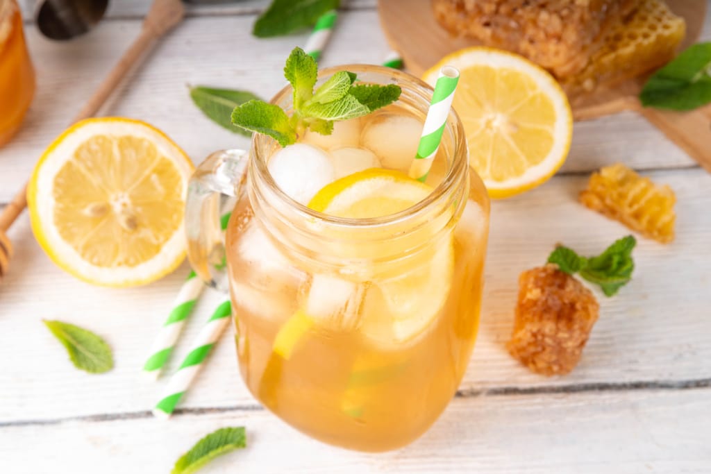 Pöydällä lasi keltaista juomaa, jossa jääpaloja ja sitruunaa. Vieressä pillejä.