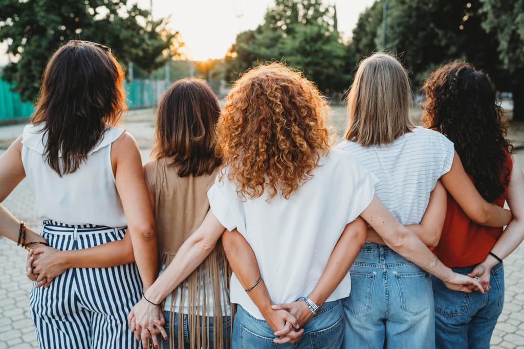 Viisi naista selin kameraan pitää toisiaan kädestä.