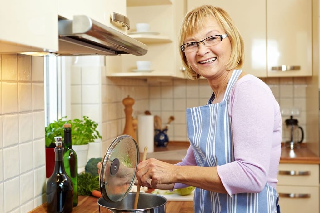 Hymyilevä keski-ikäinen nainen siniraitaisessa esiliinassa sekoittaa puulusikalla kattilassa kotikeittiössä