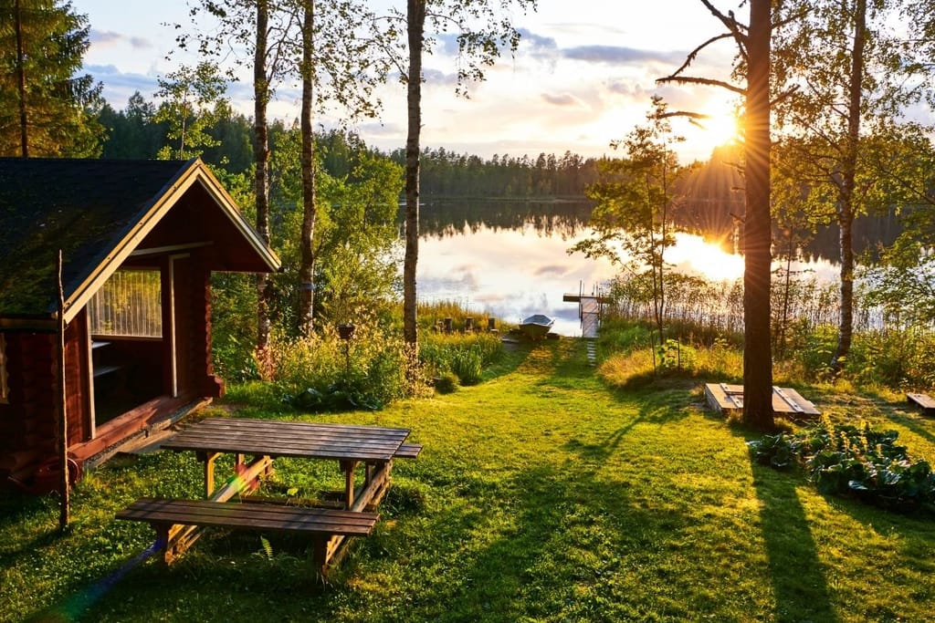 Suomalainen kesämökki järven rannalla iltaruskon valossa, edessä nurmikko ja piknikpöytä, taustalla vene ja koivut.