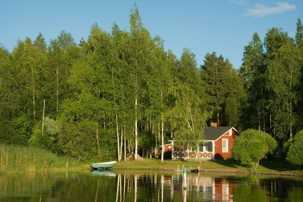 Punainen mökki järven rannalla keskellä metsää kuvaa suomalaisen mökkikesän hyvinvointivaikutuksia.