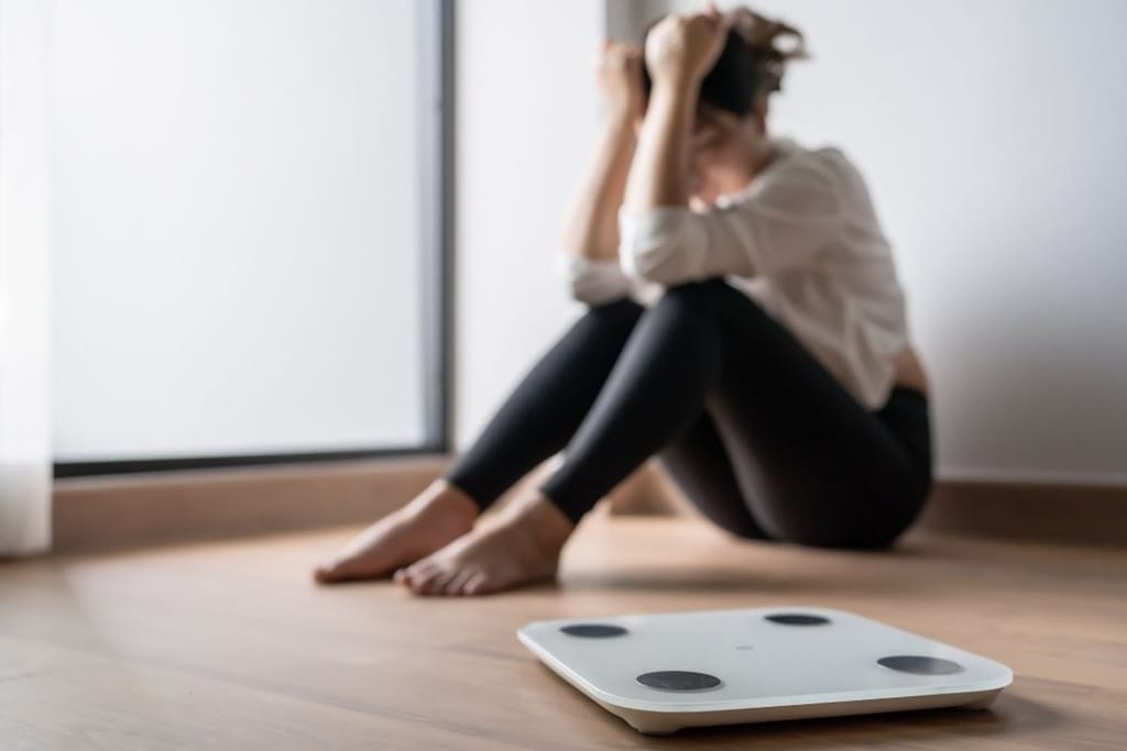 Digitaalinen henkilövaaka etualalla ja taustalla lattialla istuva turhautunut nainen epätarkkana.