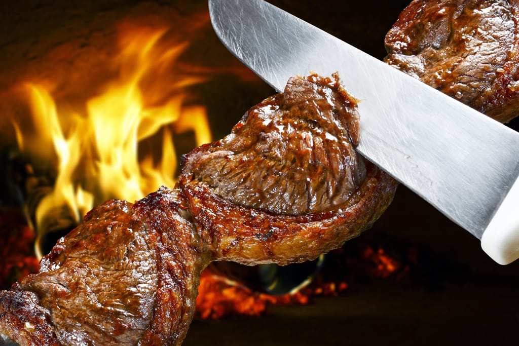 Tarjoilija leikkaa mehukasta grillattua naudanlihaa suuresta churrasco-vartaasta avotulen loimussa. Kuvassa näkyy Cavernan brasilialaistyylinen rodízio-tarjoilu, jossa lihaa tarjoillaan suoraan asiakkaiden pöytiin.