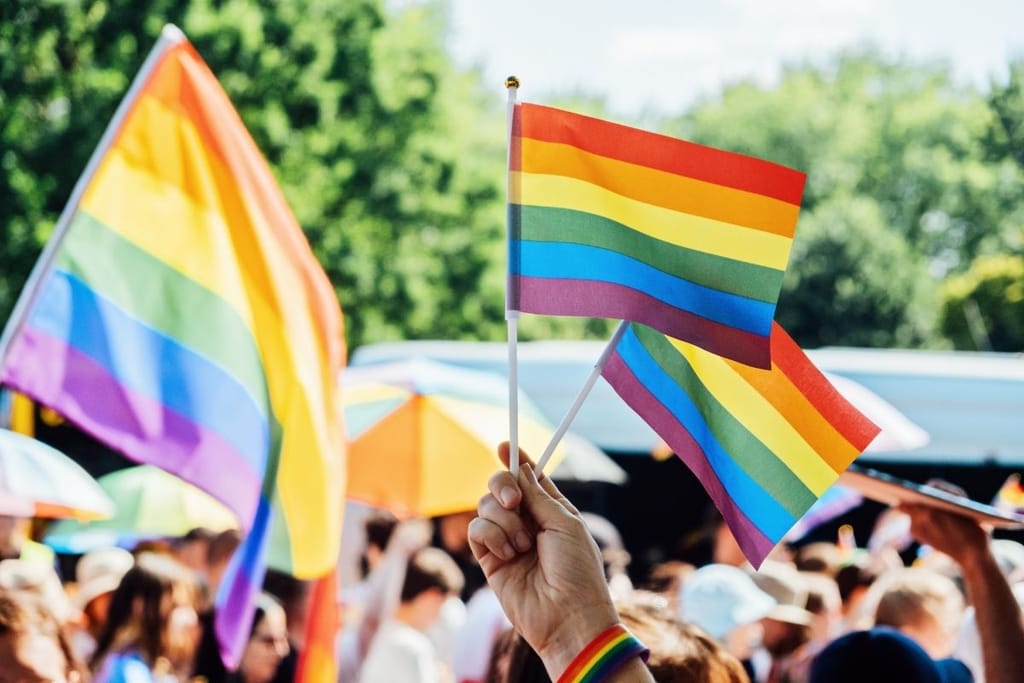 Lähikuva kädestä, joka pitää kolmea sateenkaarilippua Pride-kulkueen väkijoukossa. Liput ovat kirkkaanvärisiä ja tausta on sumea.