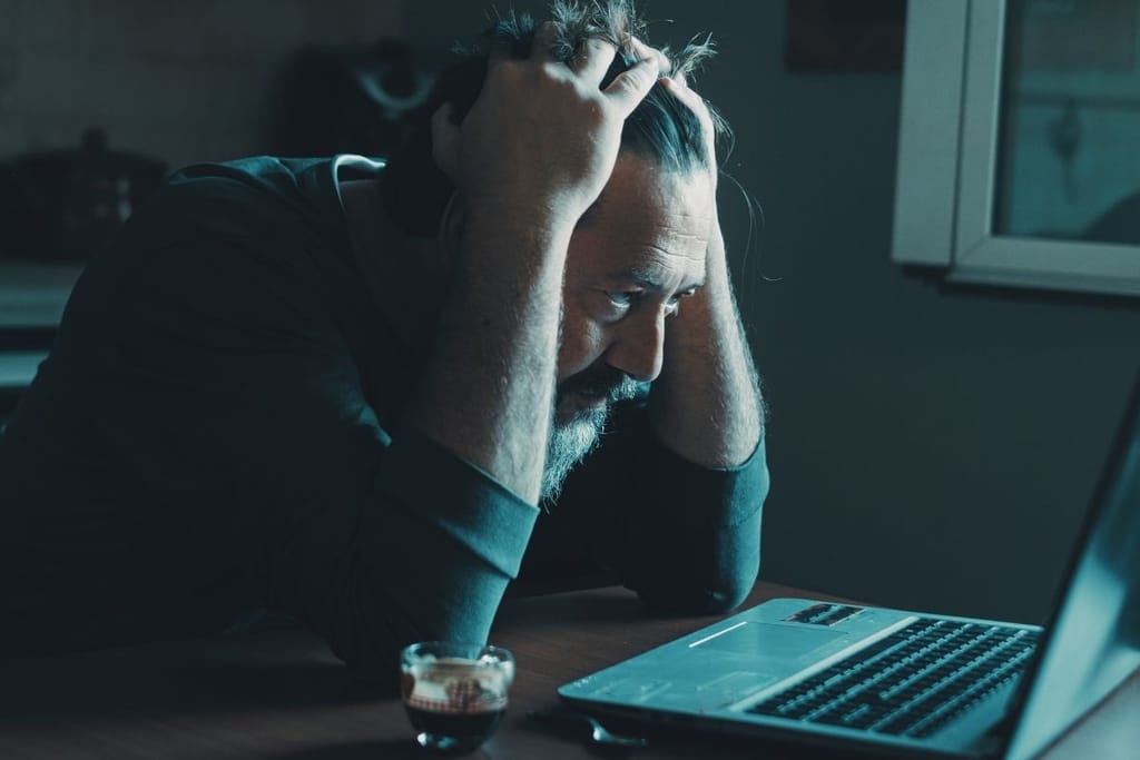 Stressaantunut ja epätoivoinen mies pitelee päätään tietokoneen ääressä hämärässä huoneessa.