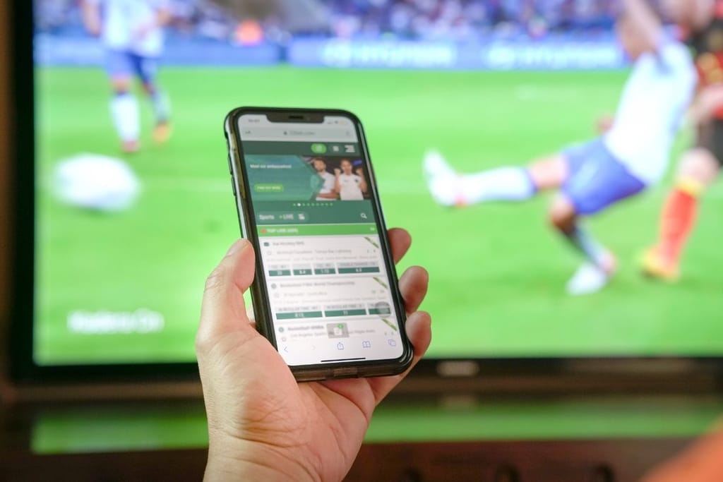 Käsi pitää älypuhelinta edessään jalkapallolähetystä näyttävän television edessä