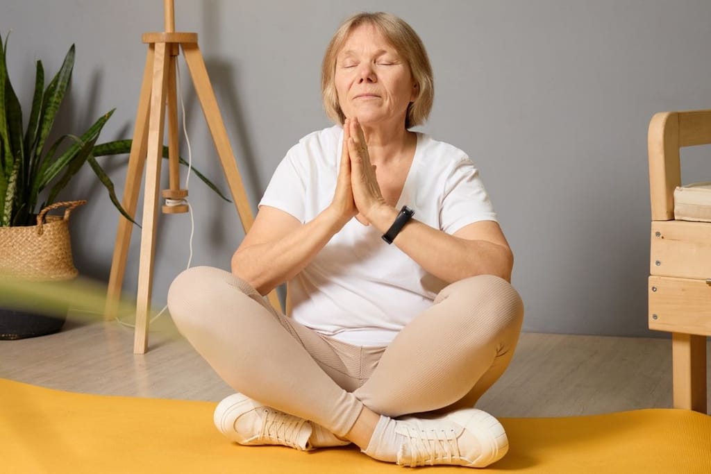 Iäkkäämpi nainen istuu ristijaloin joogamatolla silmät suljettuina, kädet rinnalla meditaatioasennossa