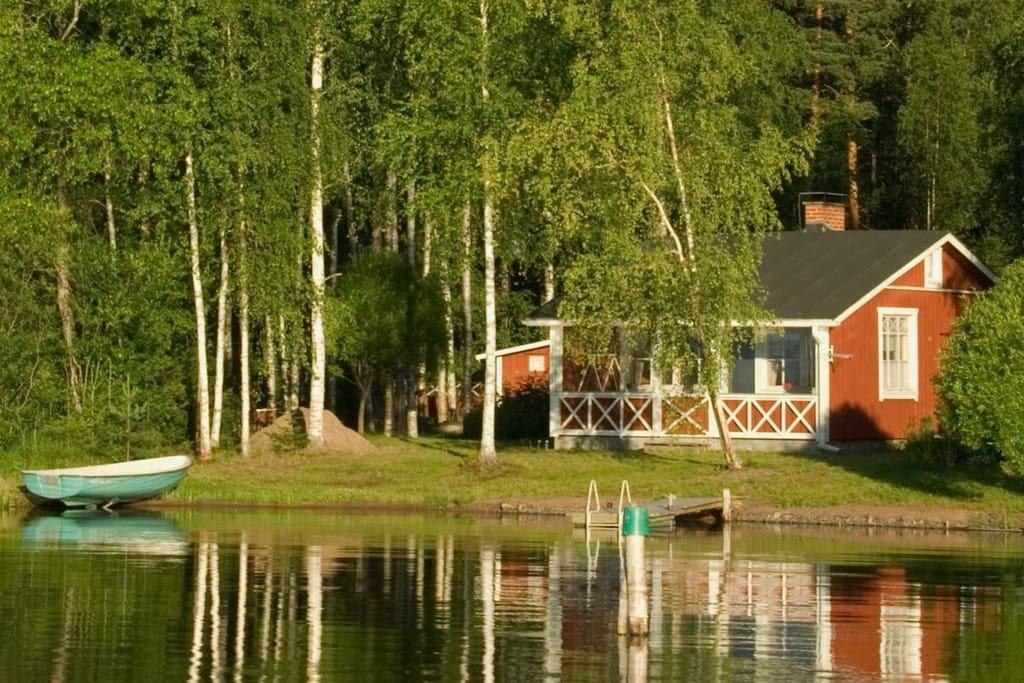 Punainen kesämökki ja vihreä soutuvene tyynen järven rannalla koivujen katveessa ilta-auringon valossa.