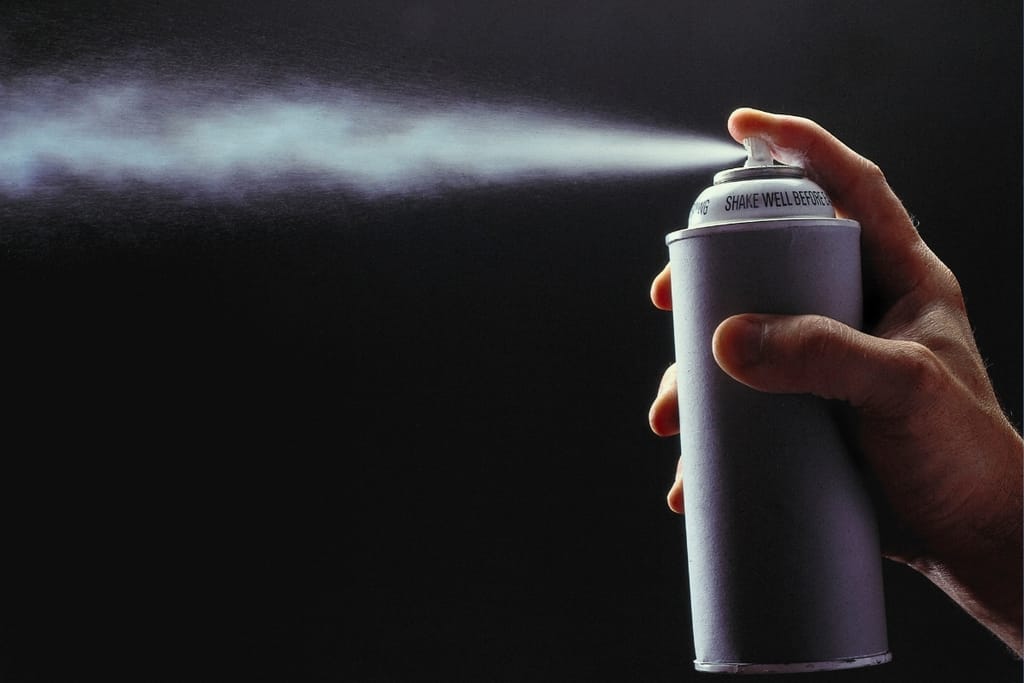 Käsi suihkuttaa paineistettua aerosolipulloa tummaa taustaa vasten.