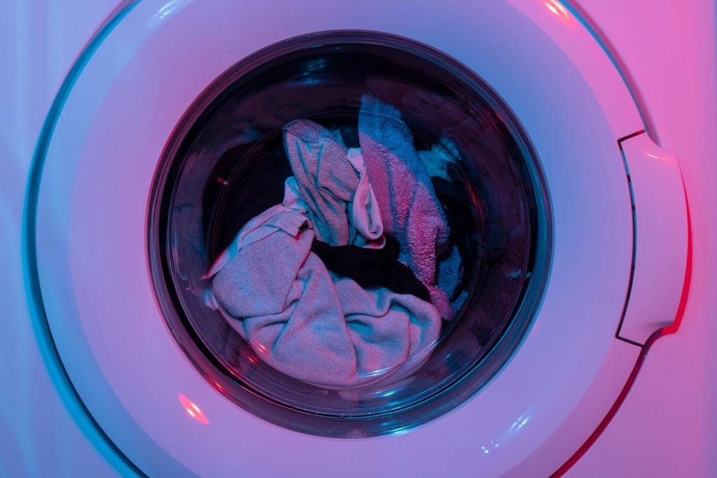 Pyykinpesukoneen lasiluukku ja täyteen ahdetut vaatteet rummussa korostettuna sinisellä ja punaisella neonvalolla.