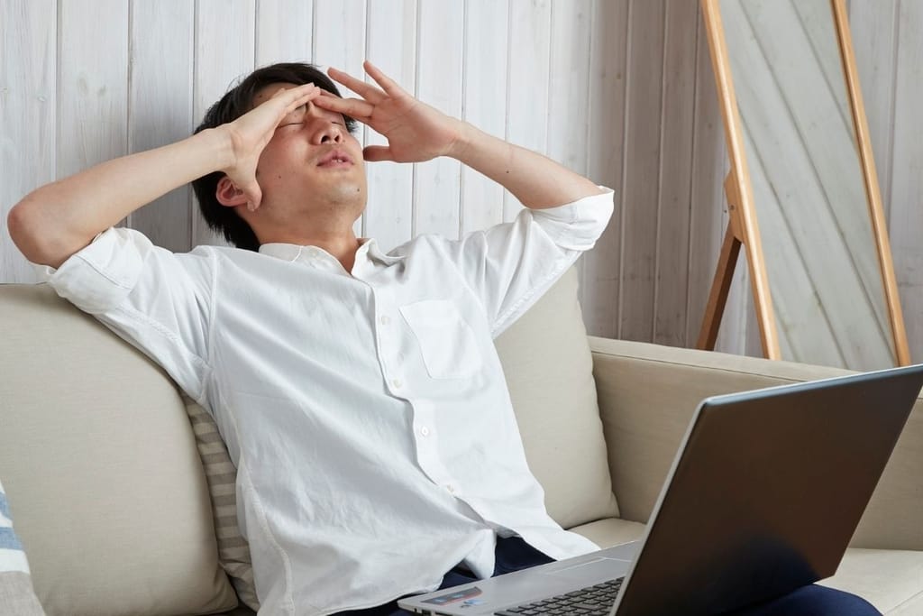 Valkoiseen kauluspaitaan pukeutunut mies istuu sohvalla kannettavan tietokoneen ääressä ja puristaa silmiään sekä otsaansa tuskaisen tai stressaantuneen ooisena.