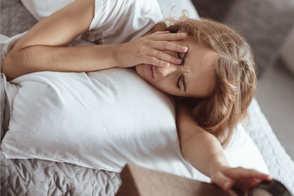 Nainen makuuhuoneessa heräämässä sängystä pää valkoisella tyynyllä ja hieromassa silmiään tukkoisuuden tai allergiaoireiden vuoksi.
