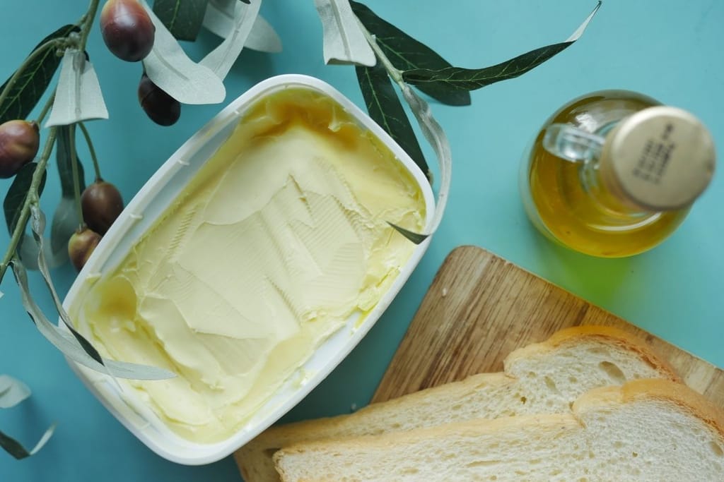 Yläviistosta otettu kuva, jossa näkyy avattu voirasia, lasinen oliiviöljypullo, vaaleita leipäviipaleita sekä oliivinoksia vaaleansinisellä taustalla.
