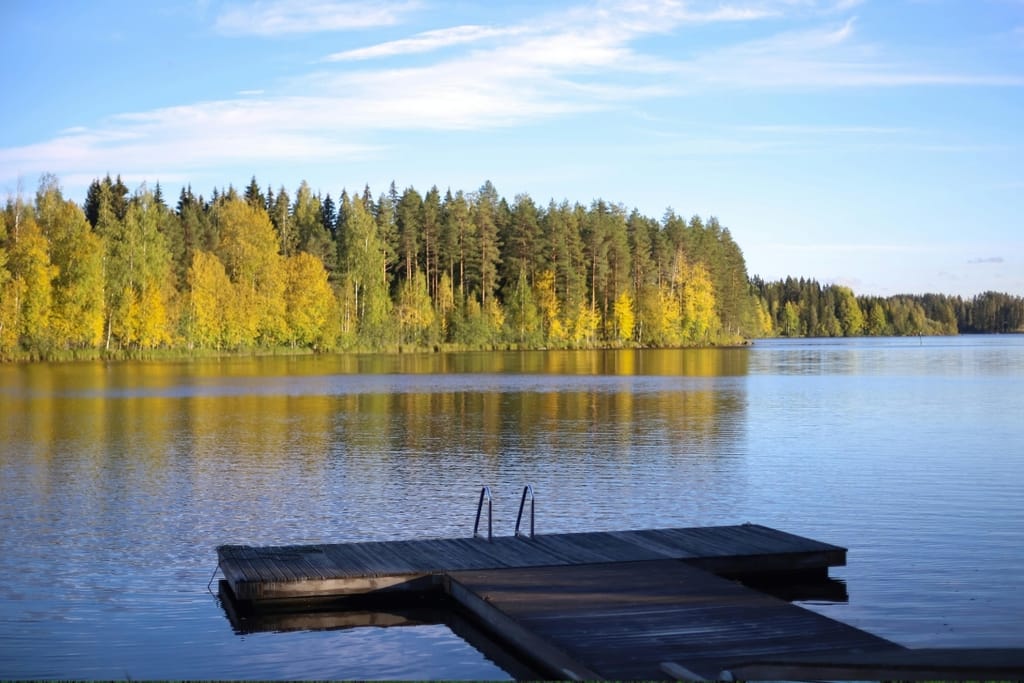 Aurinkoinen järvimaisema Suomessa, jossa laituri ja uimatikkaat, rannalla puita syksyisissä väreissä ja vihreänä.