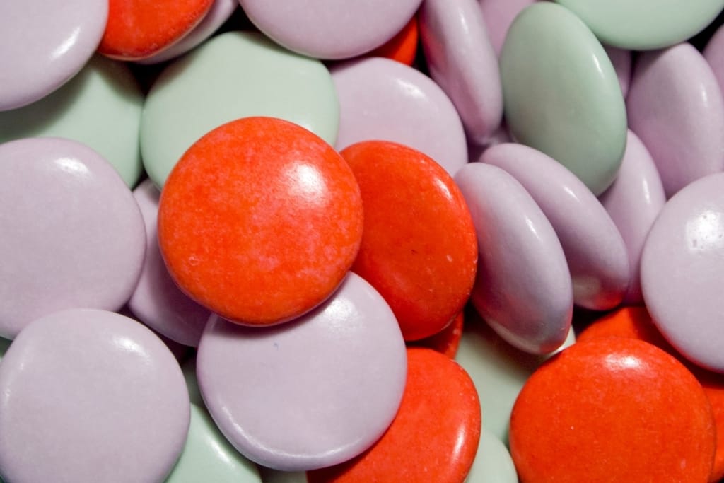 Lähikuva värikkäistä pyöreistä ja litteistä pastelinsävyisistä ning oransseista karkkirakeista ja pastilleista.