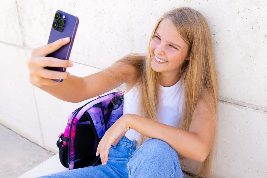 Hymyilevä pitkähiuksinen nuori tyttö ottaa selfietä puhelimellaan nojaten seinään koulureppu vieressään.