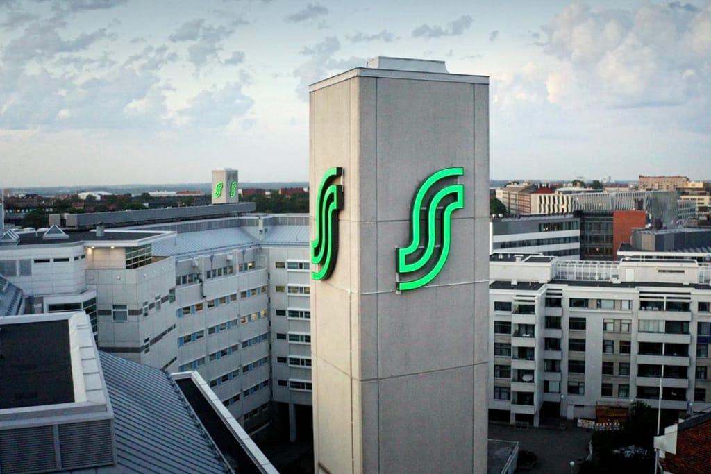 S-ryhmän valaistu vihreä S-tunnus toimistotornin seinässä, taustalla kaupungin kattoja iltavalossa.