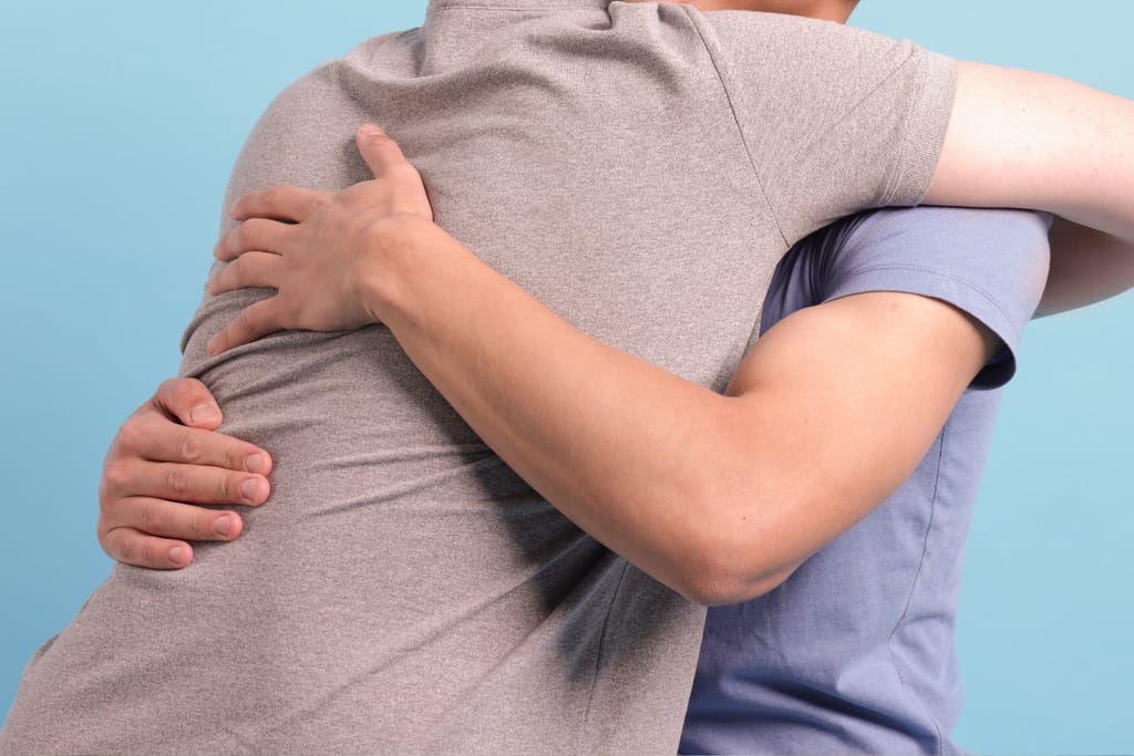 Kaksi aikuista halaa toisiaan lähikuvassa vaaleansinistä taustaa vasten.