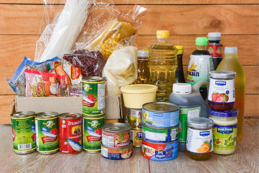 Säilyketölkkejä, pastaa, riisiä, öljyä, kastikkeita ja muita pitkään säilyviä elintarvikkeita puisella taustalla.

