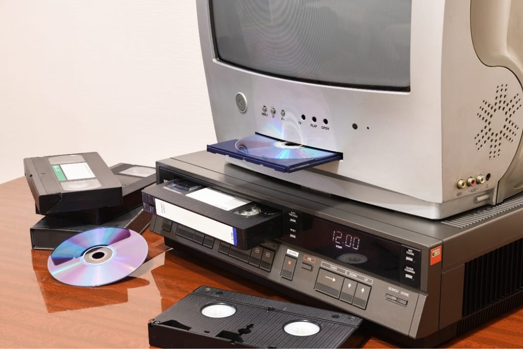 Vanha kuvaputkitelevisio, VHS-videonauhuri, videokasetteja ja CD-levyjä puisella pöydällä.