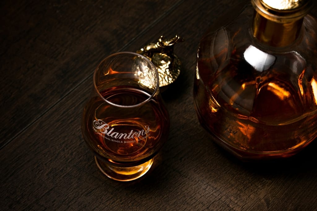 Blanton’s-logolla varustettu viskilasi ja osittain näkyvä bourbonviskipullo tummalla puupöydällä.