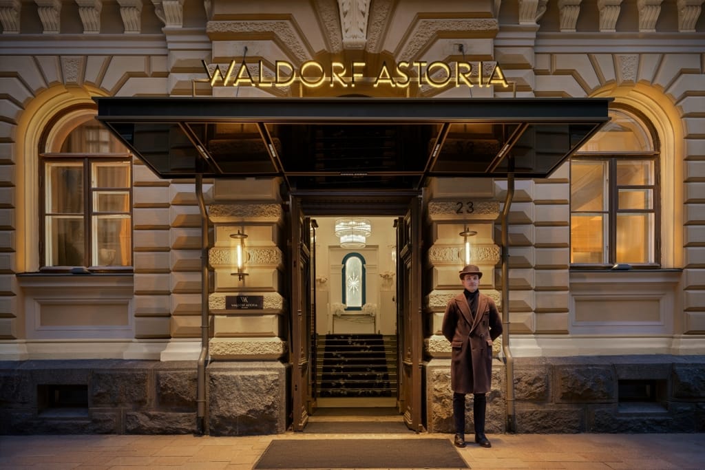 Waldorf Astoria Helsinki -luksushotellin valaistu sisäänkäynti Helsingin Kruununhaassa. Oven edessä seisoo ruskeaan takkiin pukeutunut mies.