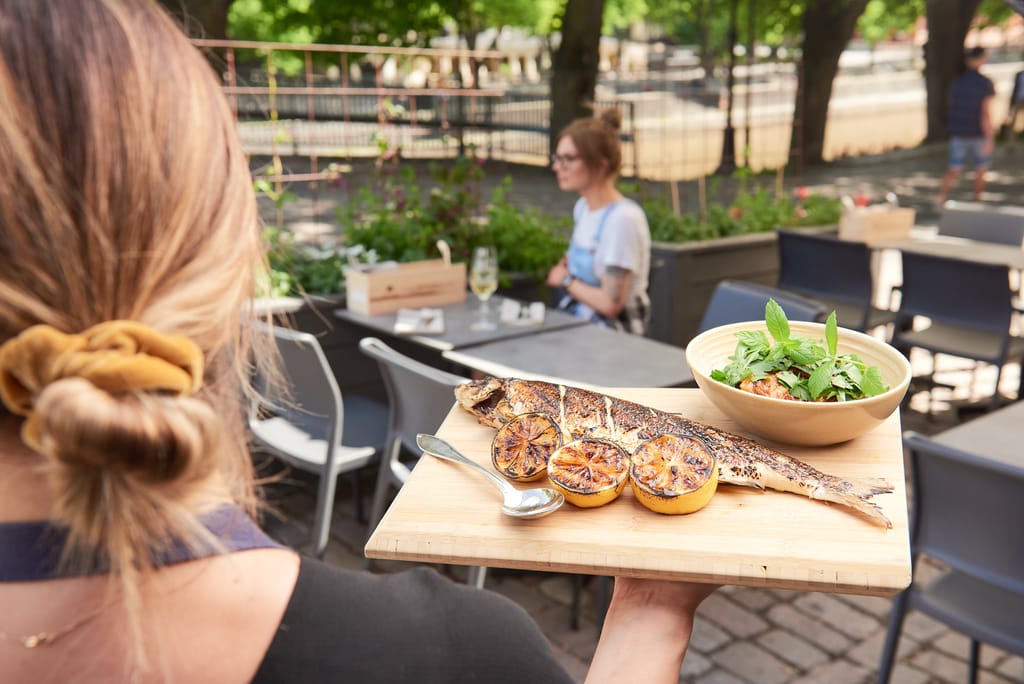 Ravintola Gustavon tarjoilija kantaa puulautasella grillattua kokonaista kalaa, paahdettuja sitruunoita ja yrttisalaattia ravintolan terassilla.