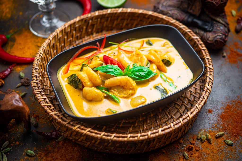 Thaimaalainen curryannos, jossa katkarapuja ja vihanneksia keltaisessa kookoskastikkeessa mustassa astiassa punotun korin päällä.