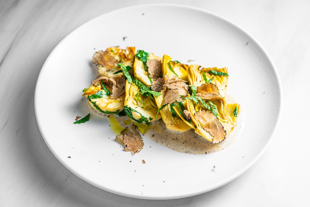 Paahdettuja artisokanpuolikkaita kermaisella kastikkeella, yrteillä ja tryffelilastuilla valkoisella lautasella.