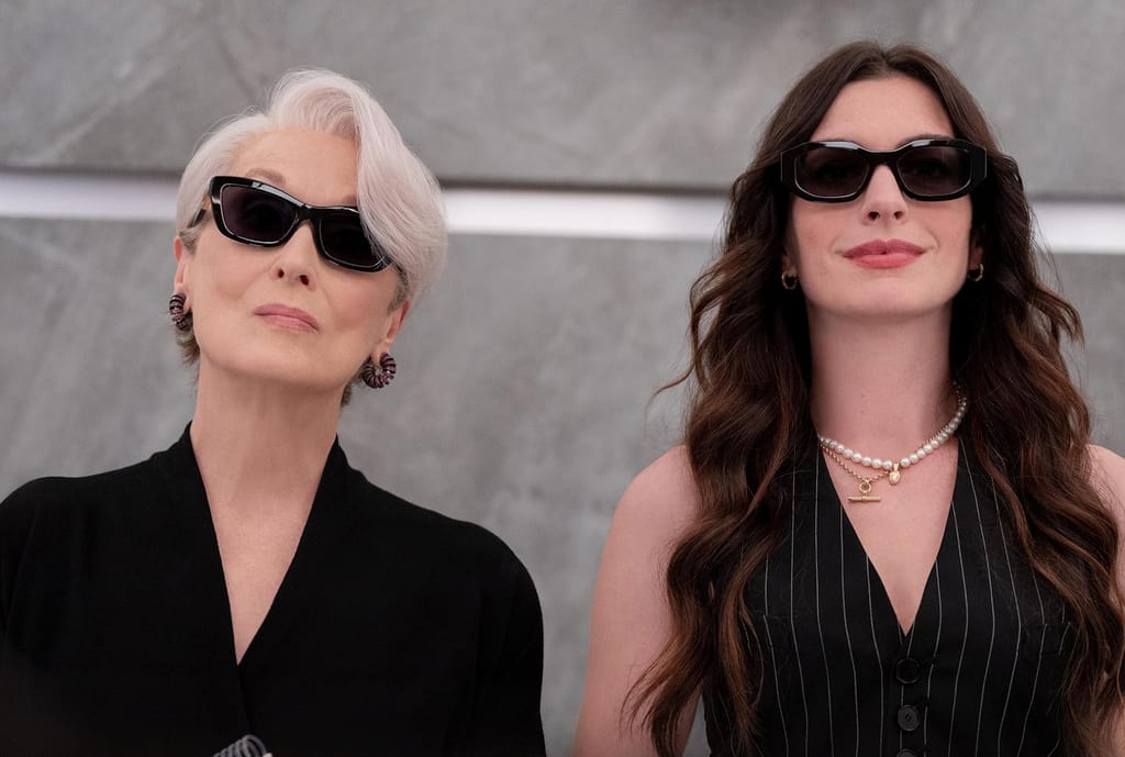 Kaksi tyylikkäästi pukeutunutta naista mustissa asuissa ja aurinkolaseissa seisoo vierekkäin – toinen harmaahiuksinen, toinen pitkähiuksinen brunetti.