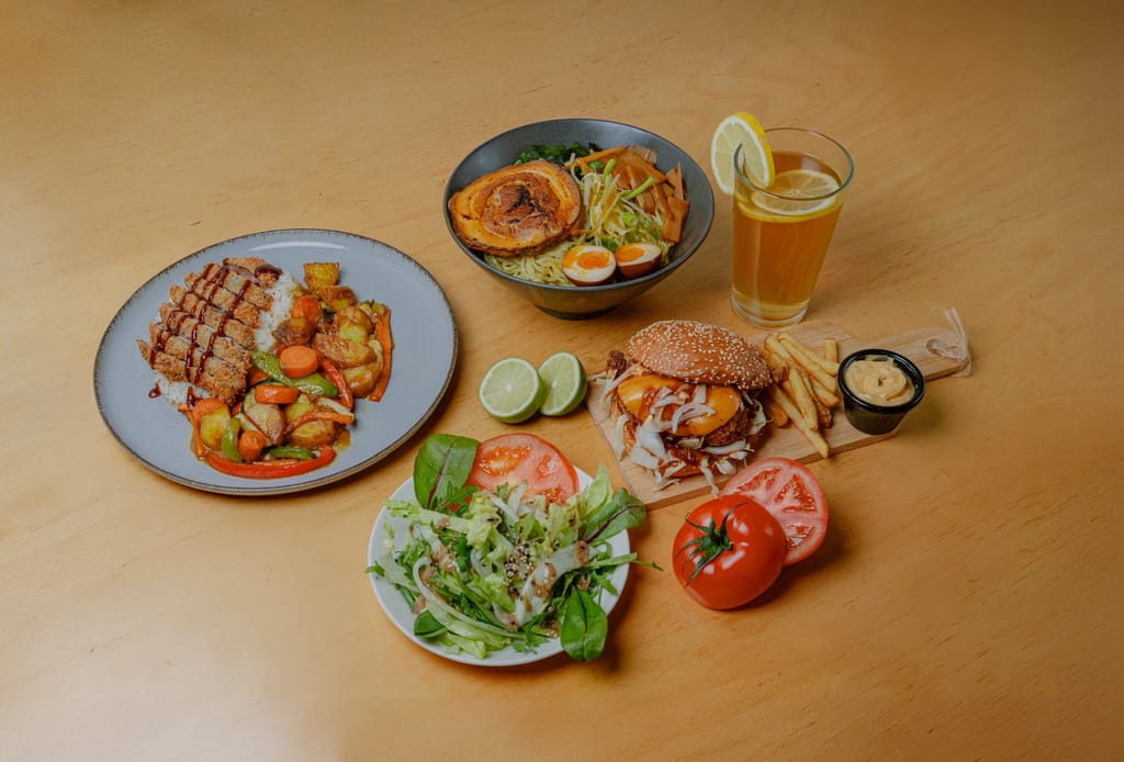 Pöydälle katettu useita ravintola-annoksia, mukana ramenkulho, hampurilainen ranskalaisilla ja dipillä, vihersalaatti sekä riisi- ja kasvisannos.