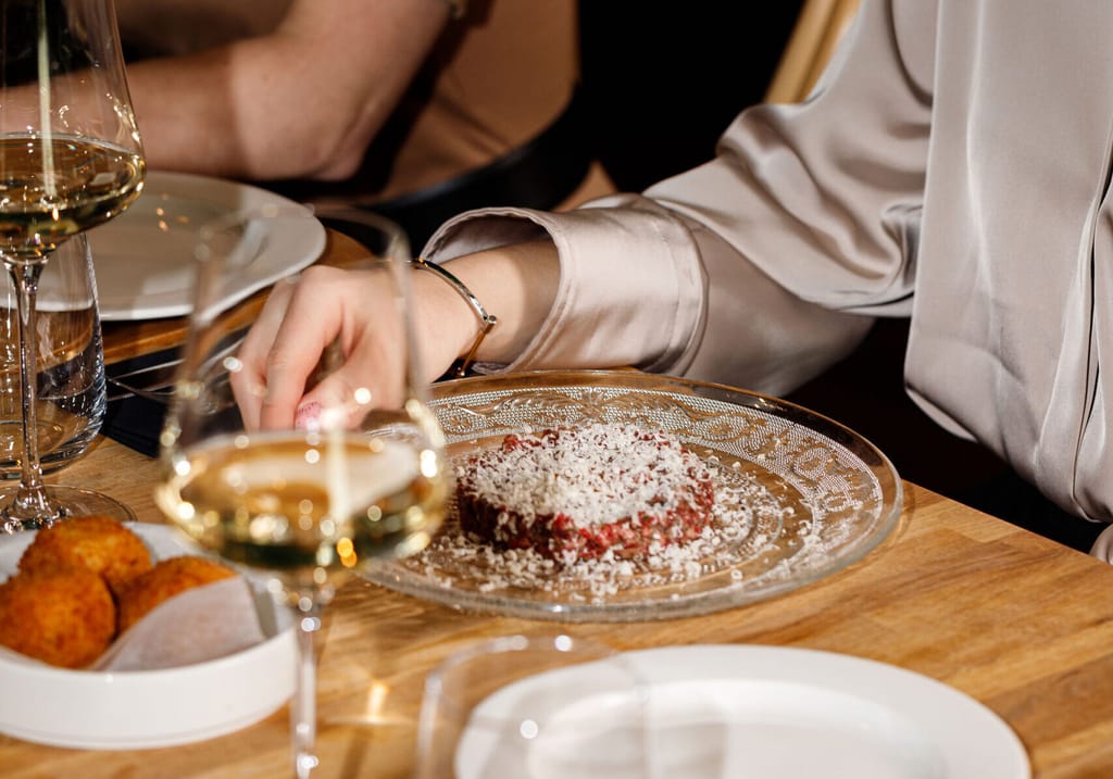 Ravintolapöydässä tarjottu annos ja viinilasit, kaksi ruokailijaa pöydän ääressä.
