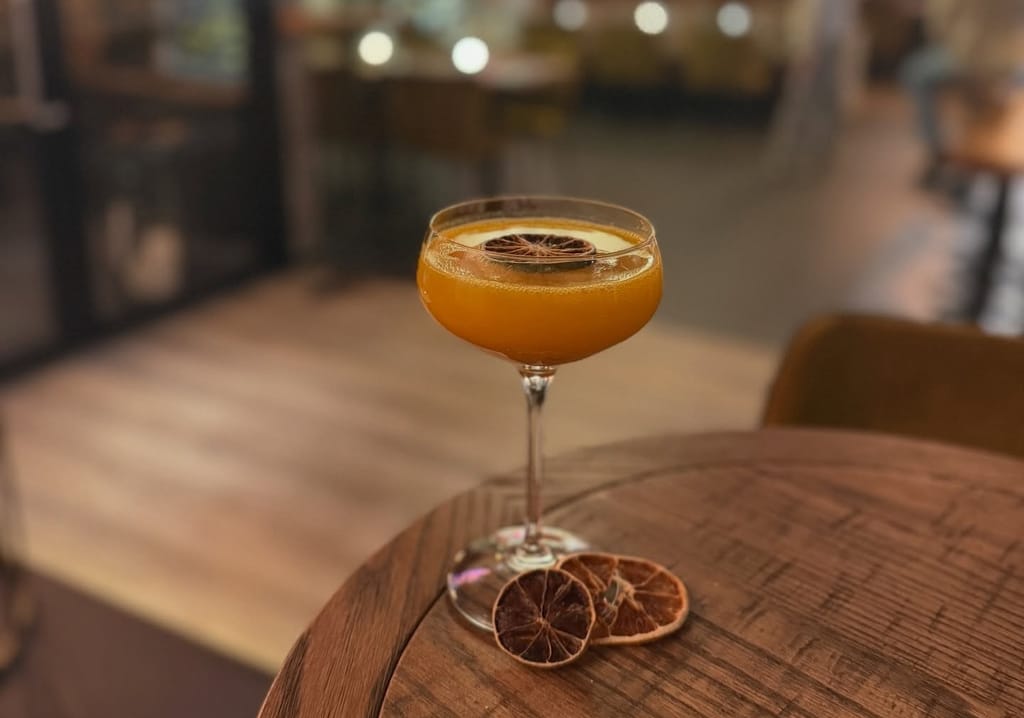 Kirkkaanoranssi cocktail jalallisessa lasissa puupöydällä, koristeena kuivattu sitrussiivu; taustalla hämärä ravintolamiljöö.