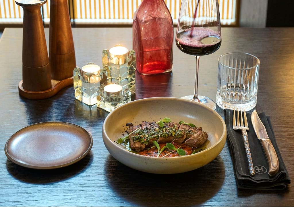Ravintola-annos, jossa lihaa ja kastiketta syvässä kulhossa, pöydällä viinilasi, kynttilöitä ja ruokailuvälineet.