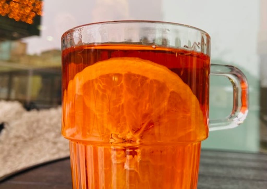 Lasimuki kuumaa teetä, jossa appelsiiniviipale, kahvilan ikkunan äärellä.