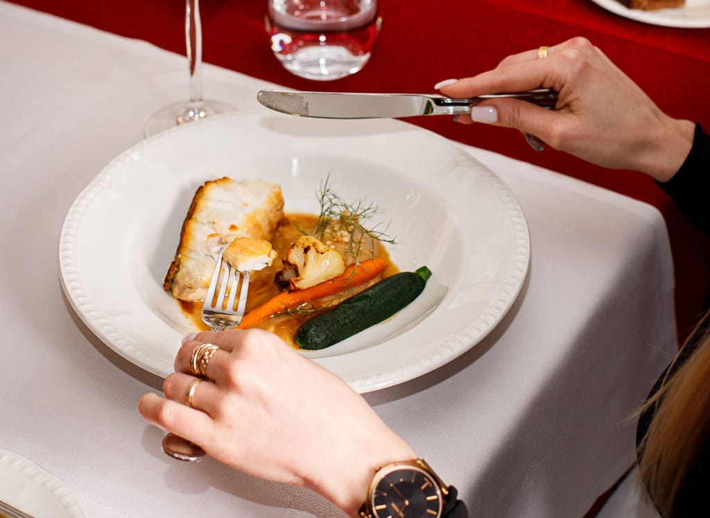 Henkilö leikkaa veitsellä ja haarukalla annosta, jossa on paistettua kalaa, kasviksia ja kastiketta valkoisella lautasella ravintolapöydässä.