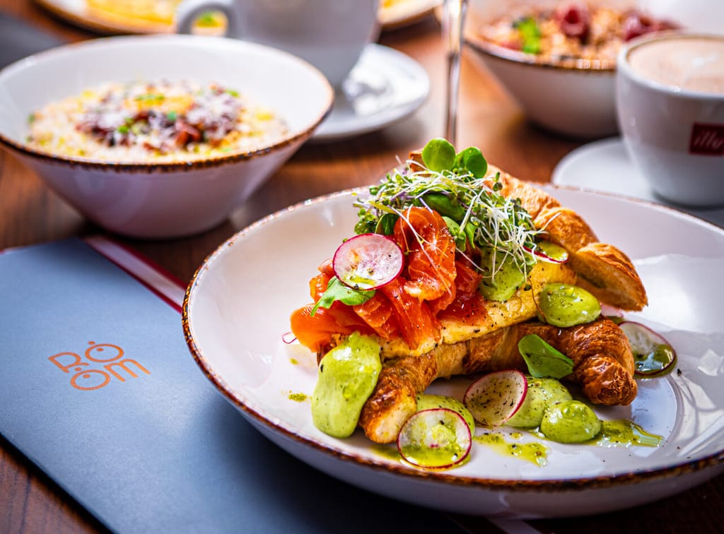 Aamiaispöytä ravintolassa: croissant lohella ja vihreällä kastikkeella, kahvikuppeja ja lasi kuohuviiniä.