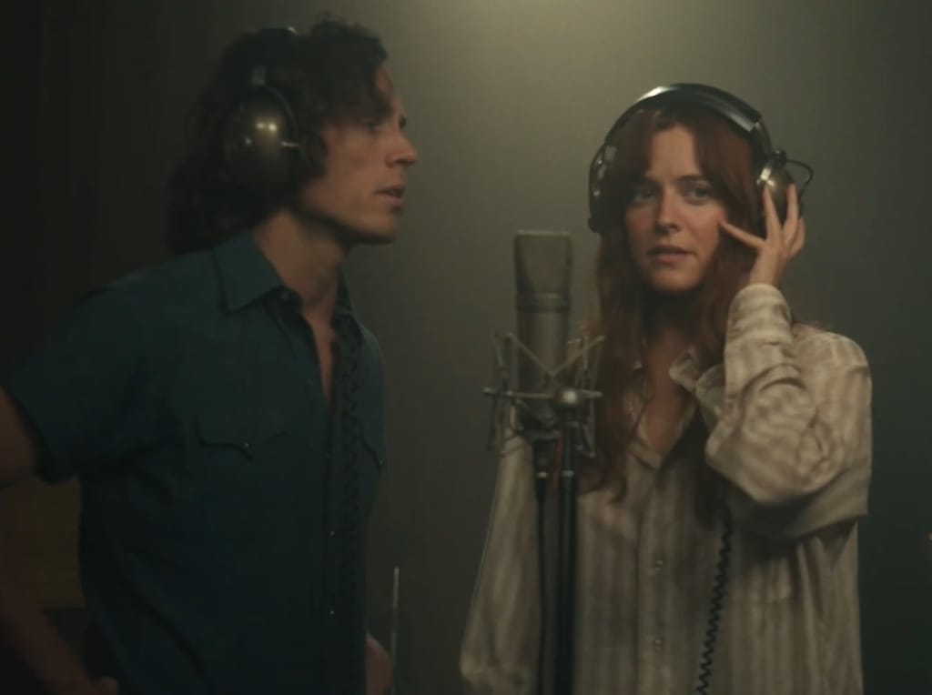 Riley Keough Sam Claflinin kanssa kuulokkeet päässä studio-olosuhteissa. Kuvassa naisella on kuulokkeet päässä ja Claflinilla myös, mutta Riley seisoo mikin edessä. 