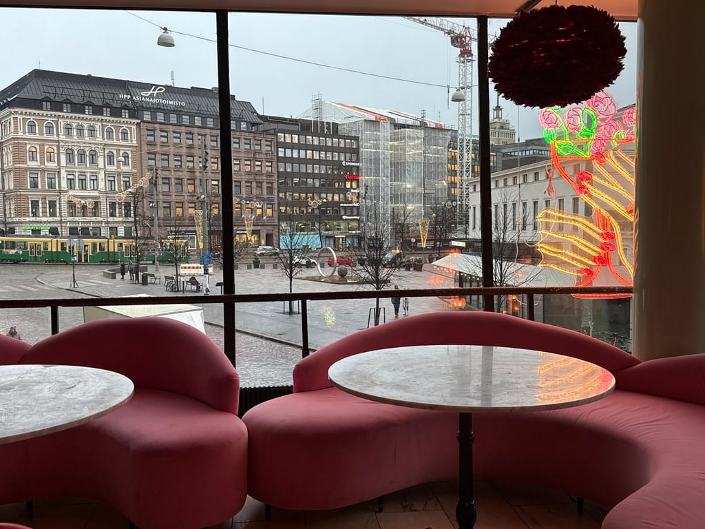 Ravintolan vaaleanpunaiset nojatuolit ja pyöreät pöydät suurten ikkunoiden äärellä, näkymä kaupunkiaukiolle.