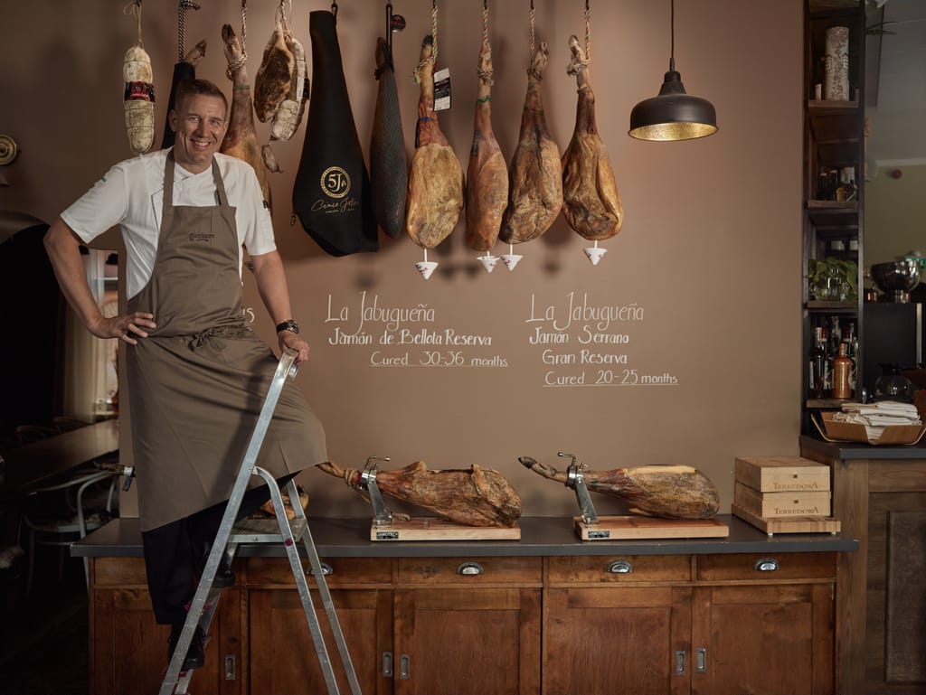 Kokki seisoo tikkailla ravintolassa, jossa seinällä roikkuu useita ilmakuivattuja kinkkuja.