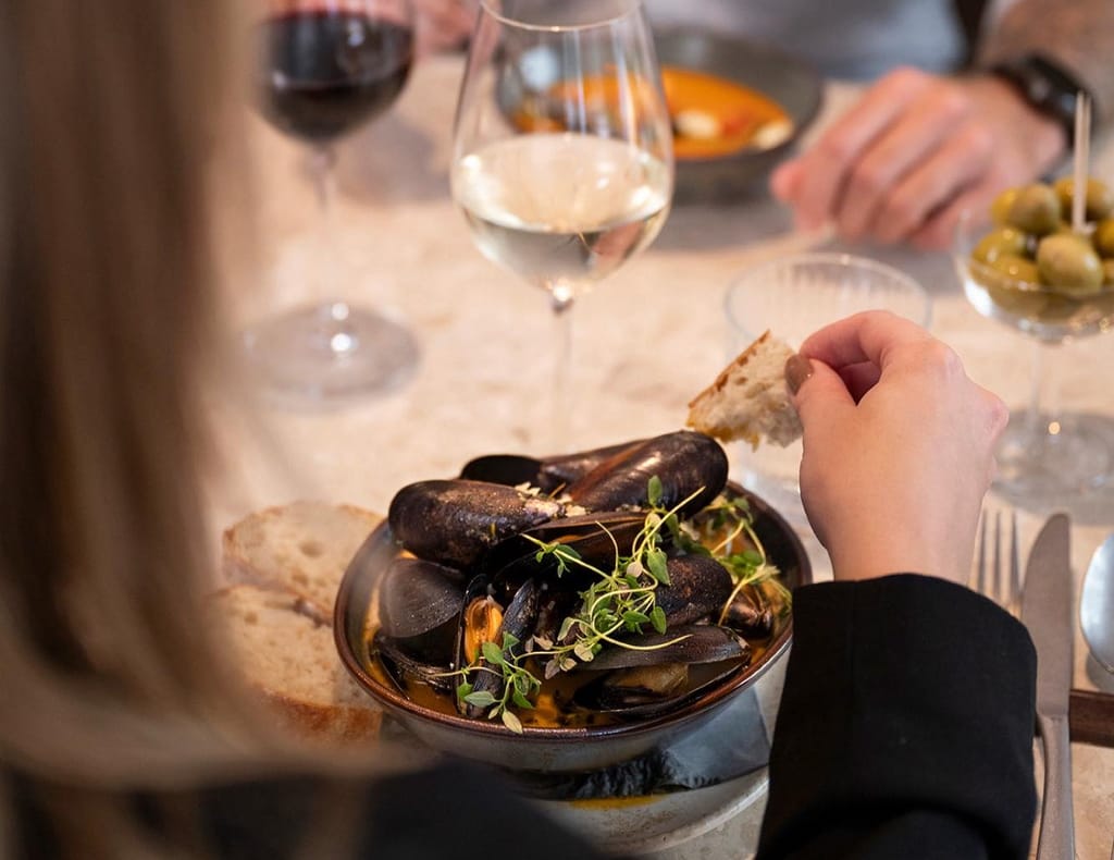 Höyryävä simpukka-annos kulhossa, vieressä leipää ja viinilasi; käsi kastaa leipää liemeen.