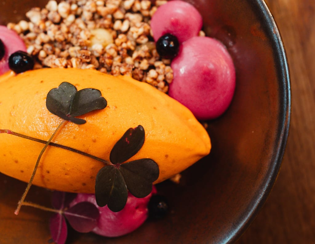 Jälkiruoka-annos, jossa oranssi mousse, marjoja, murua ja syötävä kasvinlehti tummalla lautasella.